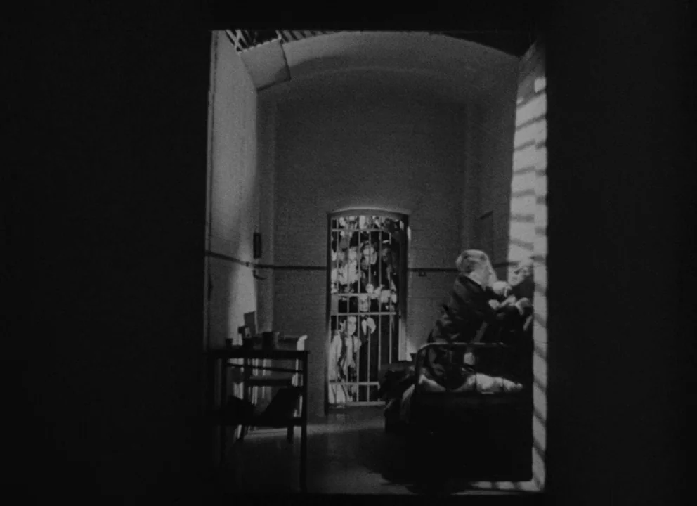 Dead of Night (1945) movie still: wide — A stark stone room features a cot and a barred entry door. A man sits on the cot while figures cluster…