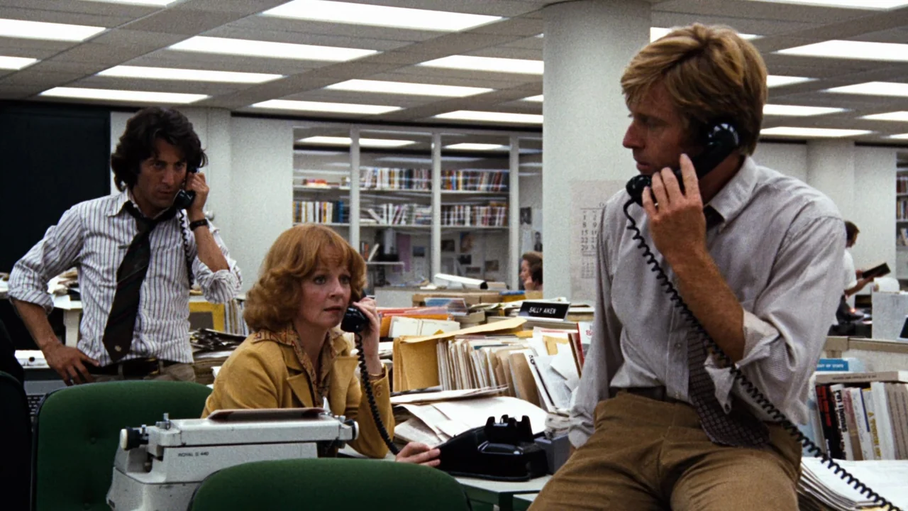 All the President's Men (1976) movie still: medium — An open-plan 1970s newsroom features cluttered desks, rows of fluorescent lights, and stacked…