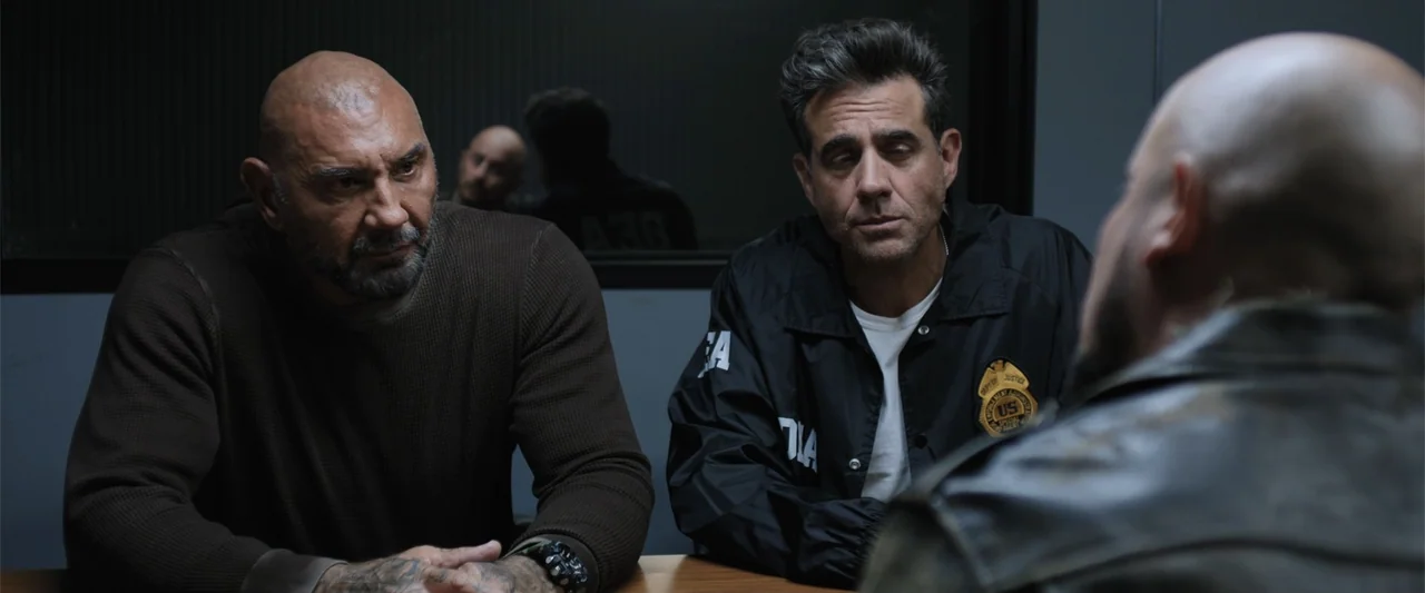 Trap House (2025) movie still: medium — A dimly lit room contains a table where three men engage in conversation. A bald man and a law enforcement…