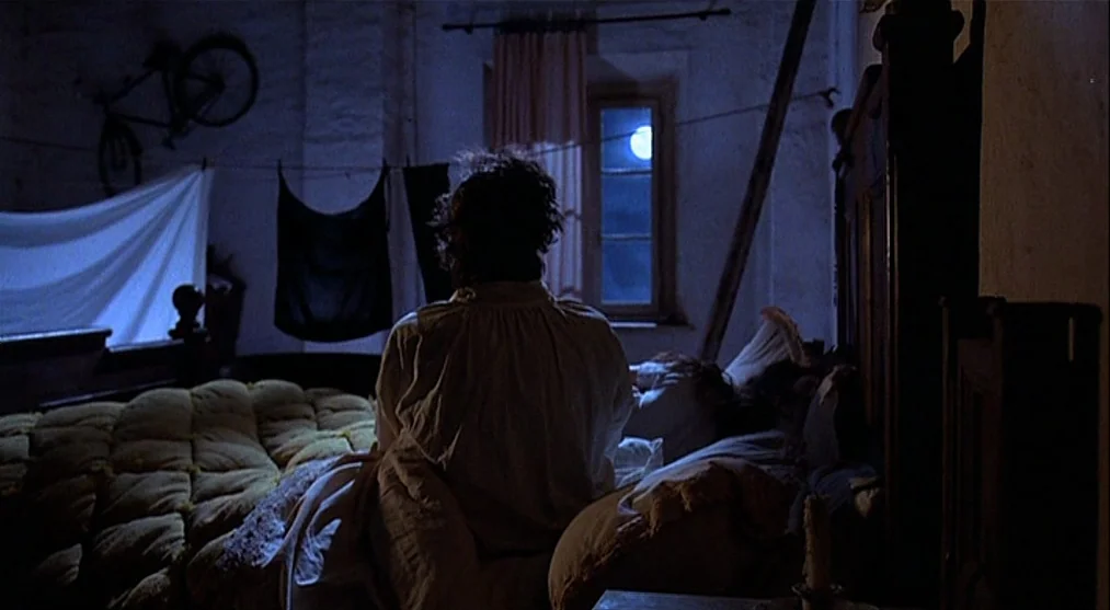 The Voice of the Moon (1990) movie still: medium — A dark, sparsely furnished room contains a bed with a thick quilted blanket. A person sits…