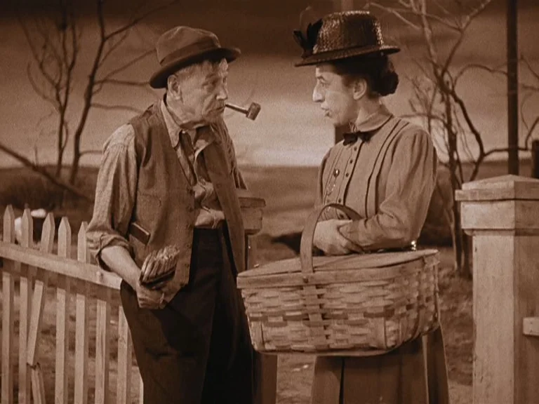 The Wizard Of Oz (1939) movie still: medium — A rural setting featuring a wooden picket fence against a simplified, painted stage background. An…