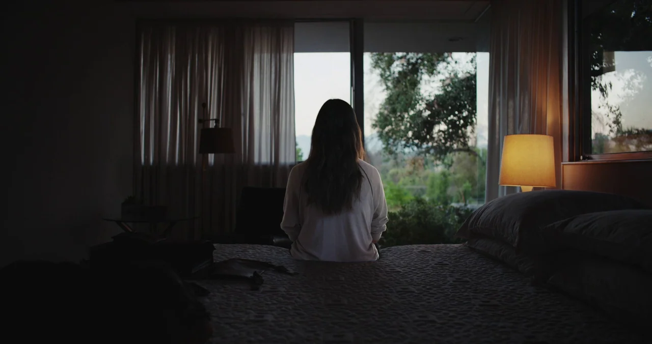 Violet (2021) (2021) movie still: medium — A modern bedroom features a large glass wall looking out onto trees at dusk. A woman sits on the edge of…
