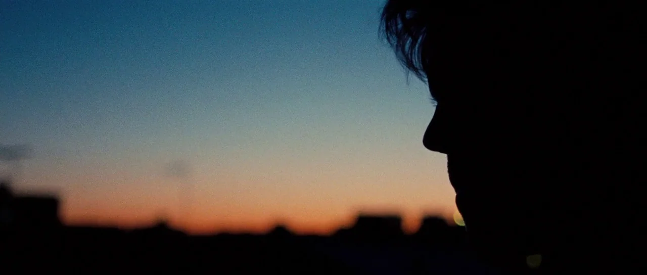 On The Road (2012) movie still: closeup — An outdoor rooftop setting at dusk overlooks an indistinct urban skyline silhouette. A person in profile…