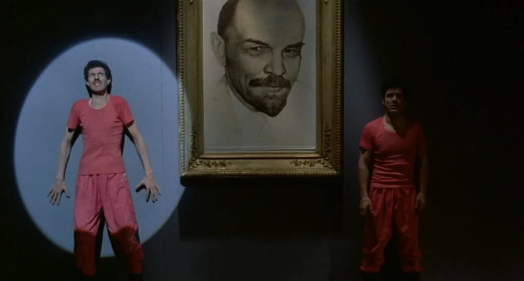 Dr. Goldfoot and the Girl Bombs (1966) movie still: fullbody — A dark, dimly lit wall displays a large framed portrait of Vladimir Lenin. Two men in…
