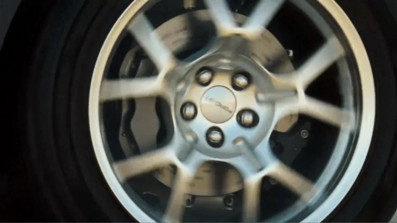 Redline (2007) movie still: closeup — A silver alloy car wheel rotates quickly against a blurry dark road surface. The rim features a central…