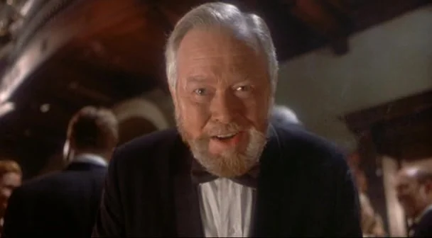 Boxing Helena (1993) movie still: closeup — A dimly lit ballroom with figures blurred in the background. An older man in a tuxedo leans forward…