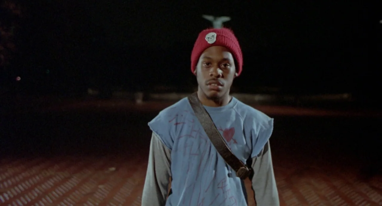 Ms. 45 (1981) movie still: medium — A dark, empty brick-paved area at night illuminated by distant ambient street lighting. A young man stands alone…