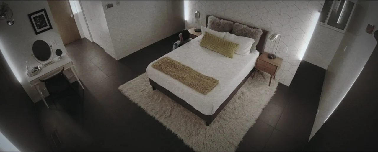 Hostages (2017) movie still: wide — A modern hotel room features a plush bed centered on a white shag rug. A person sits on the far edge of the bed…