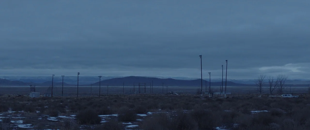 Nomadland (2020) movie still: establishing — A vast, barren desert plain stretches toward distant, snow-dusted mountains under a heavy overcast sky.…