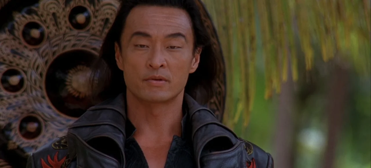 Mortal Kombat (2021) movie still: medium — A man stands outdoors with lush green palm leaves visible in the blurred background. The subject faces…