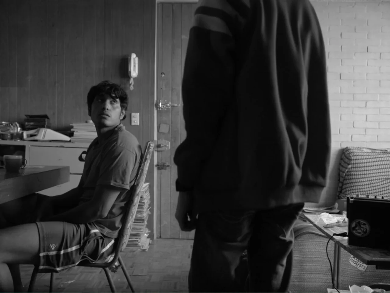 Güeros (2014) movie still: medium — A cluttered residential space features wood-paneled walls and stacks of books. One man sits in a chair looking…