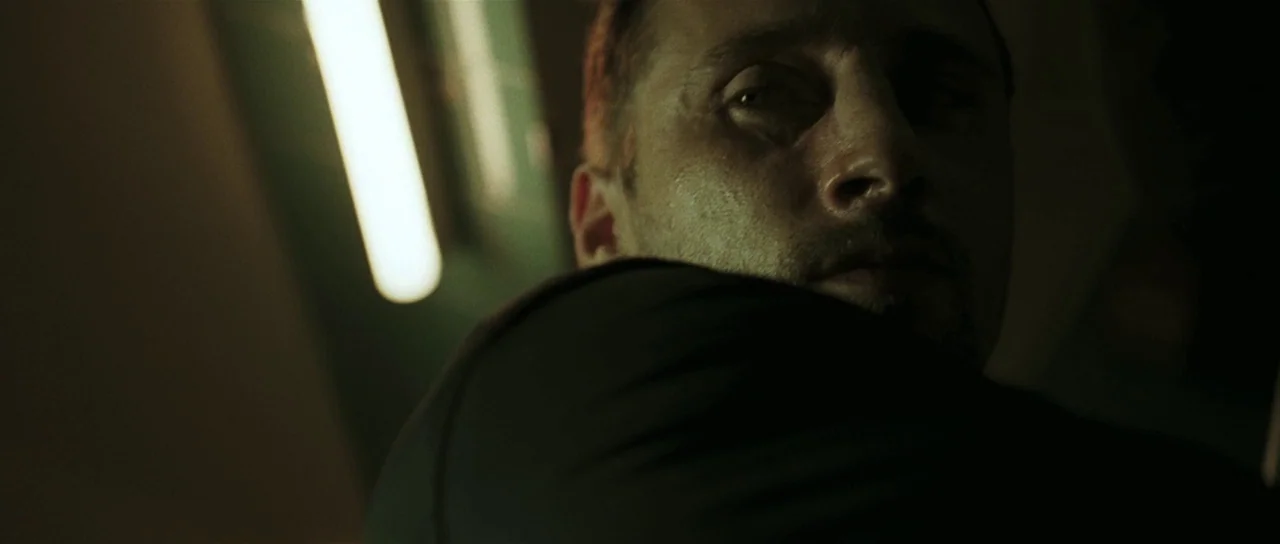 Bullhead (2011) movie still: closeup — An interior space is illuminated by a harsh, vertical fluorescent light source. A man stares at the camera…