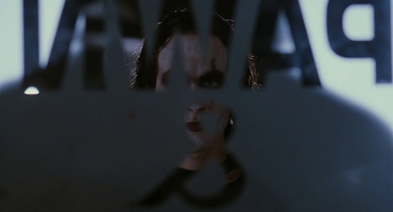The Crow (1994) movie still: closeup — A man peers through a shaped hole in a dark reflective metal sign. White lettering on the sign is visible out…