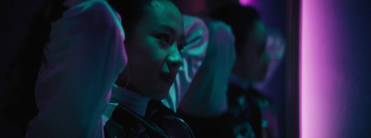 Dead Pigs (2018) movie still: closeup — A dimly lit room with a vertical neon strip casting magenta light. A woman is adjusting her hair in front of…