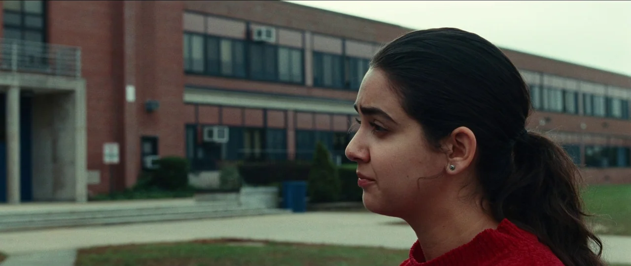 Bad Education (2019) movie still: medium — A brick school building stands in the background under a pale overcast sky. A young woman in a red…
