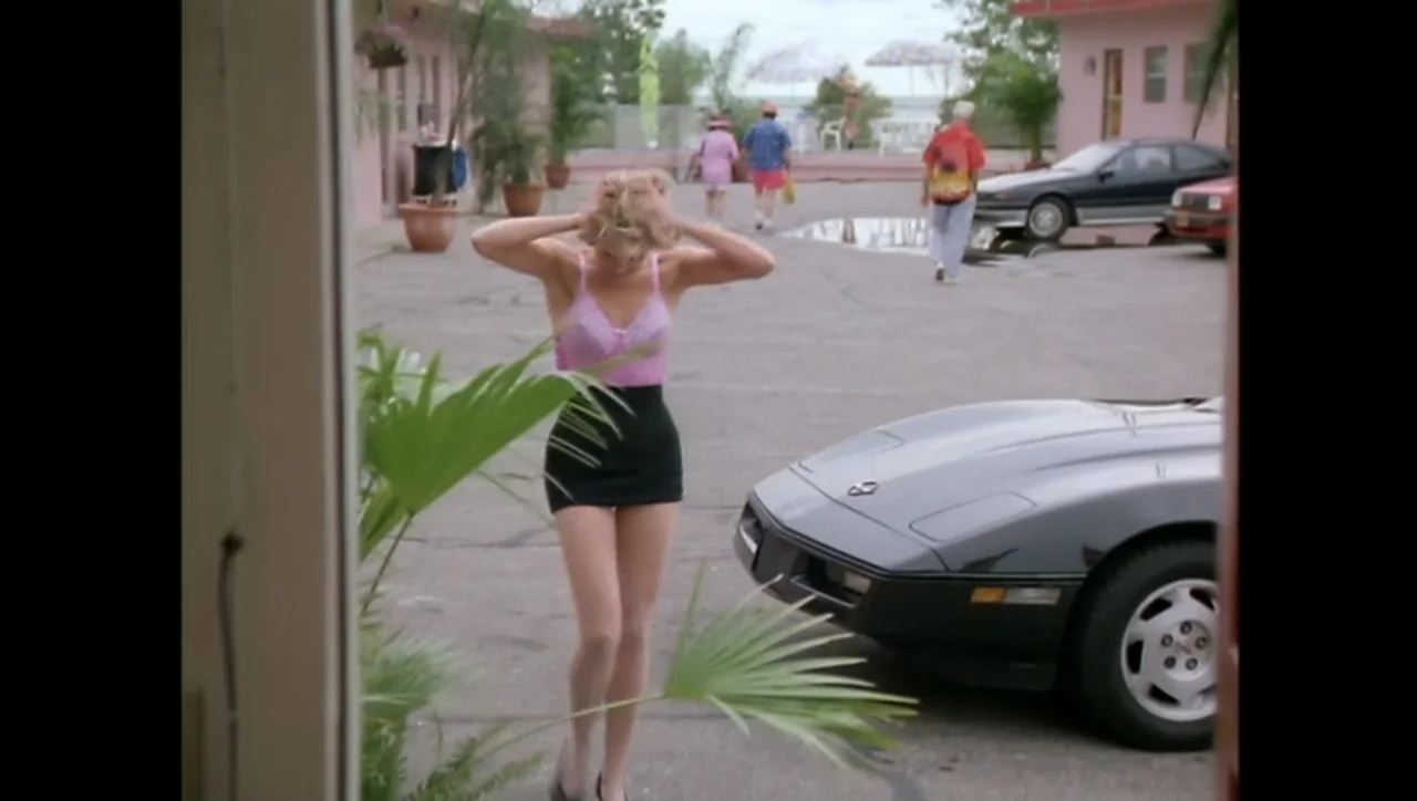 The Awakening (1995) movie still: fullbody — A pink motel parking lot features a parked black sports car in the foreground. A woman stands in the…