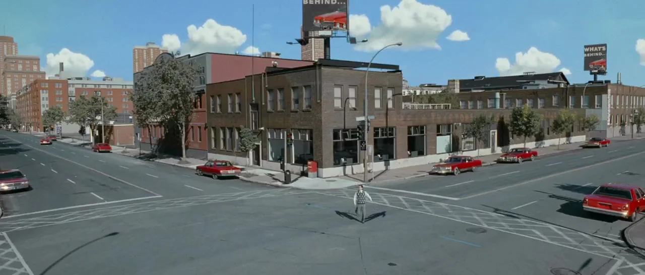 Mr. Nobody (2009) movie still: wide — A quiet urban street intersection with brick buildings and multiple parked red sedans. A man in a sweater vest…
