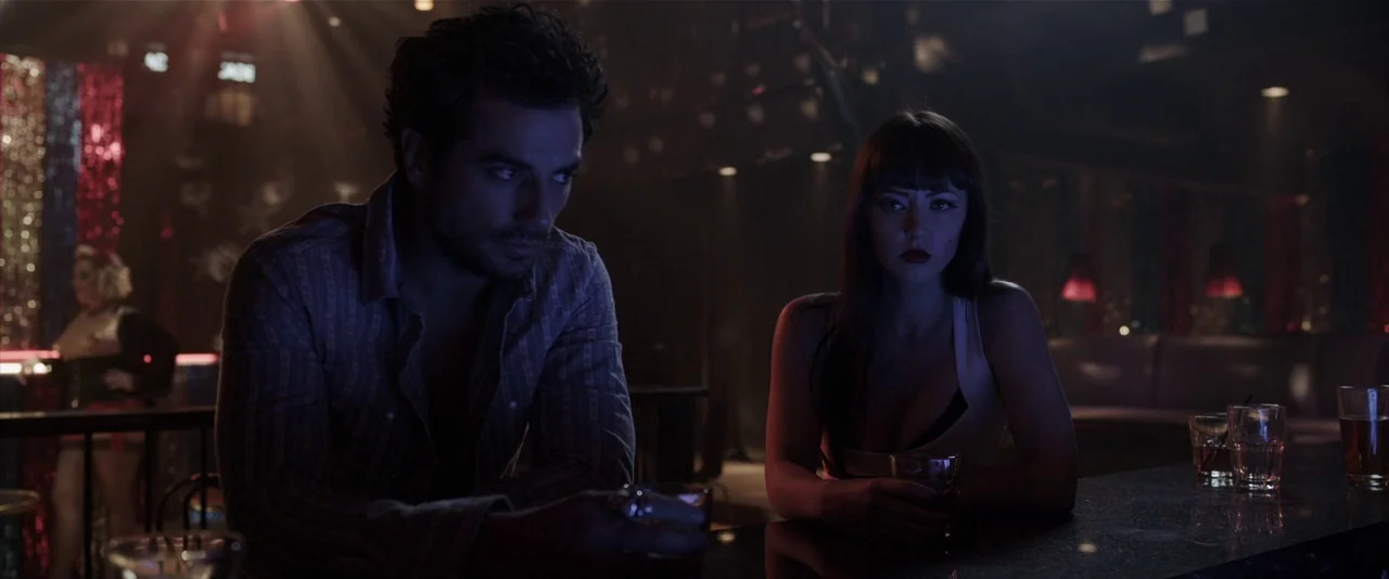 American Mary (2012) movie still: medium — A dimly lit bar features tinsel curtains and dark furniture with low visibility. A man and a woman sit…