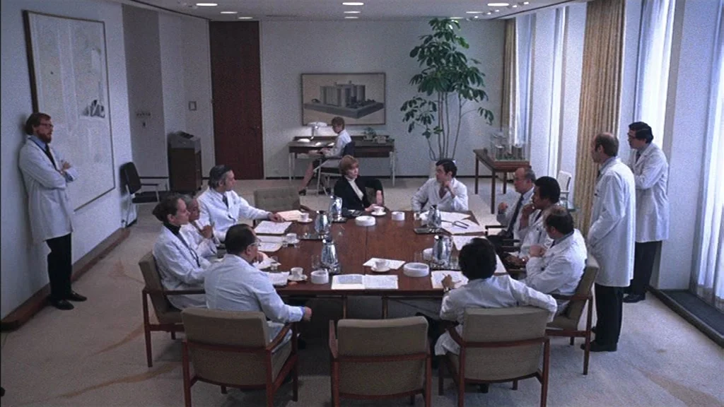 The Exorcist (1973) movie still: wide — A spacious, sterile room features a large central table surrounded by many people. Several individuals wear…
