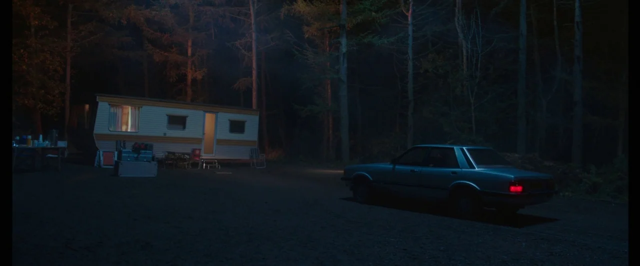 Censor (2021) movie still: wide — A secluded wooded area at night features a small stationary trailer and a sedan. The car sits idling on a dark…
