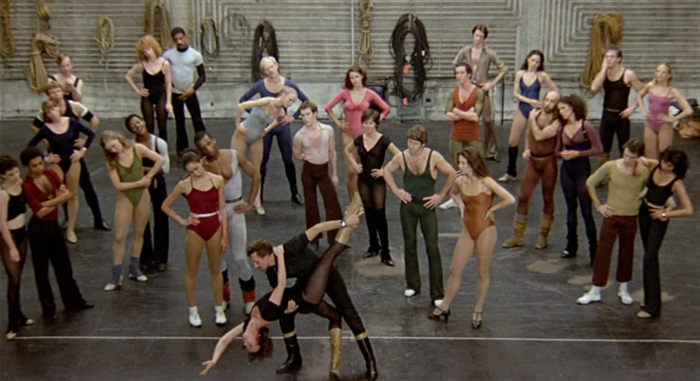 All That Jazz (1979) movie still: wide — A spacious dance studio features a black floor and gray wall with coiled ropes. A large group of dancers in…