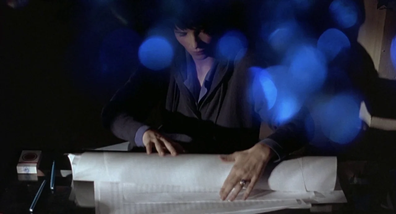 Three Colours: Blue (1993) movie still: medium — A dimly lit workspace featuring a dark desk surface cluttered with a cigarette pack. A person in a…