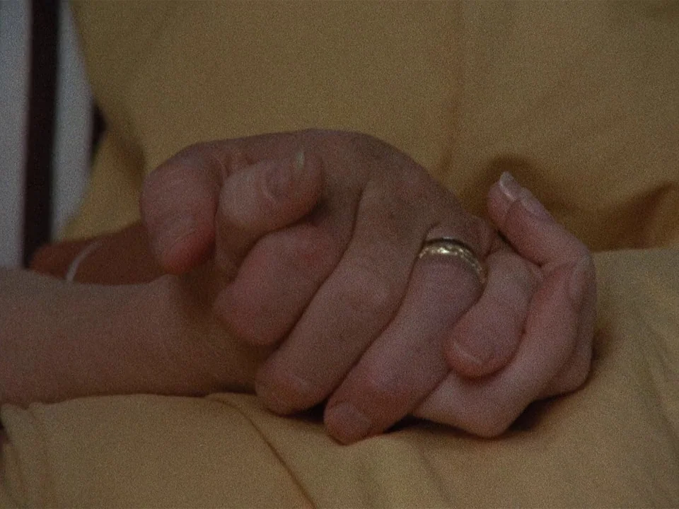 Scenes from a Marriage (1973) movie still: closeup — Hands rest on top of a draped, mustard-colored fabric surface indoors. A simple gold band is…