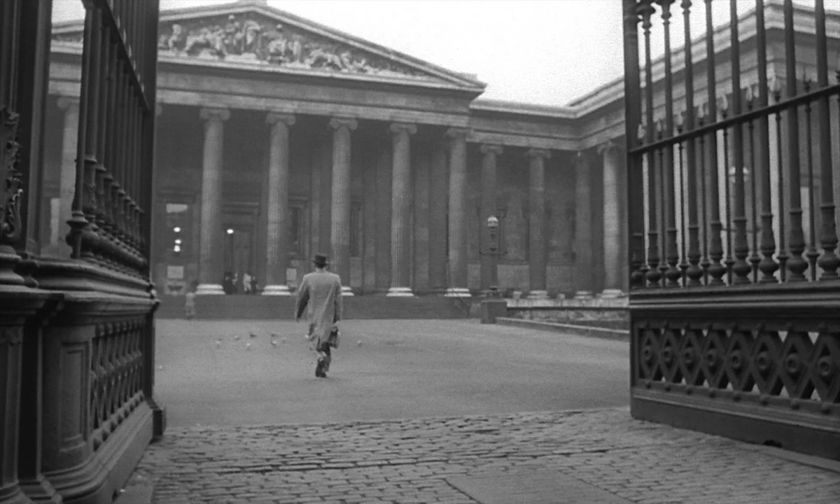 Night Of The Demon (1957) movie still: wide — A stone courtyard leads to the massive columns of the British Museum entrance facade. A lone man in a…