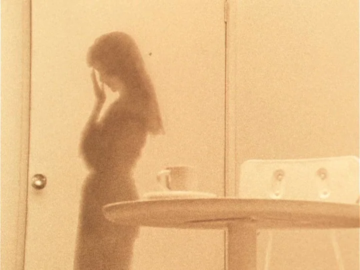 The Woman with Two Heads (1977) movie still: medium — A room with a plain wall and a round table featuring a single cup. A woman stands in profile…