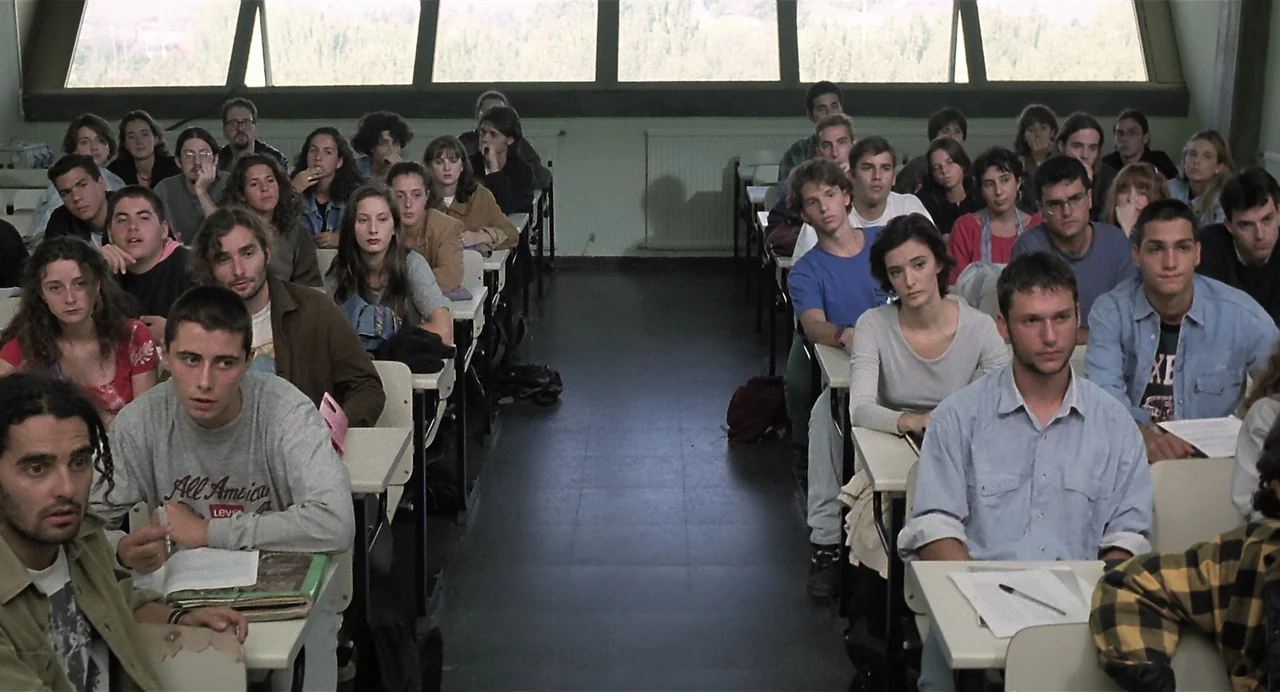 Thesis (1996) movie still: wide — A crowded university lecture hall features rows of desks facing a large windowed wall. Numerous students sit in…
