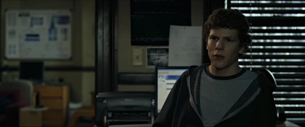 The Social Network movie still: medium — A cluttered room contains a desk, office equipment, and posters on the wall. A young man sits in front of…