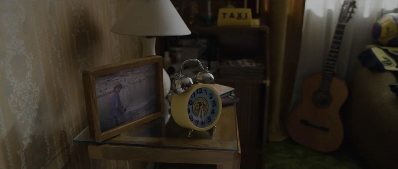 Mirreyes vs Godínez (2019) movie still: closeup — A glass-topped side table in a room contains a yellow alarm clock and picture frame. The…