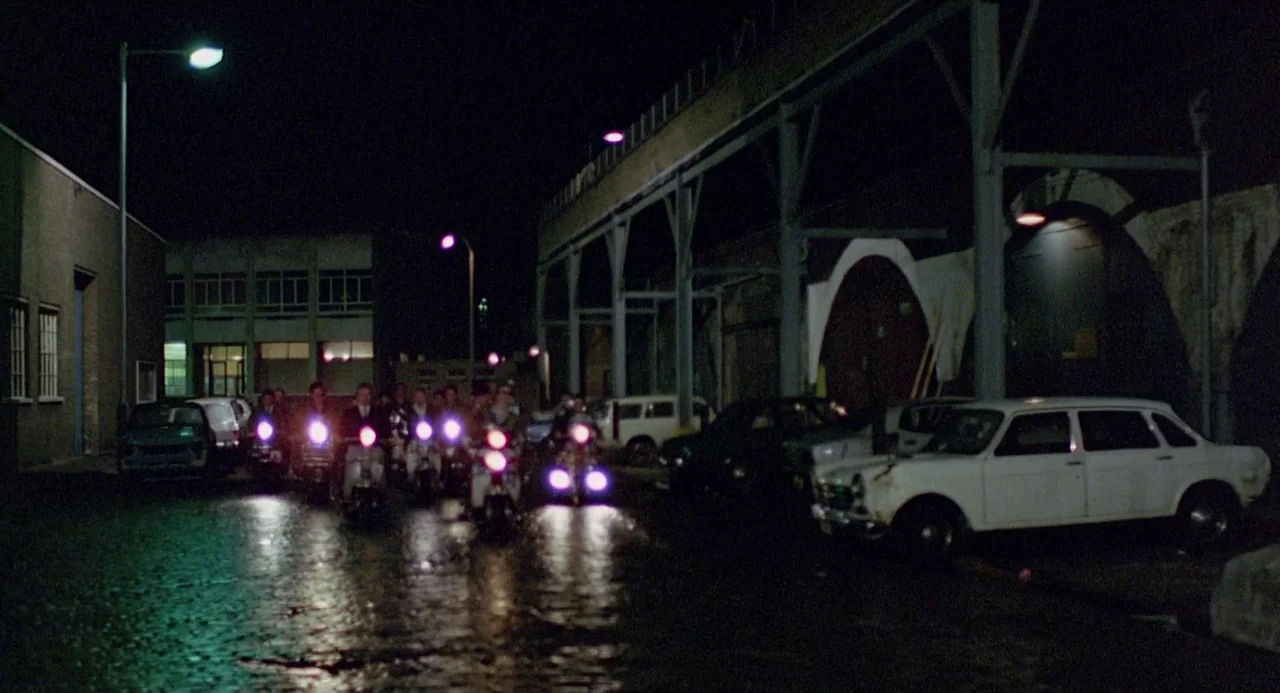 Quadrophenia (1979) movie still: wide — An urban industrial area at night with wet ground and parked vehicles along the side. A group of people…