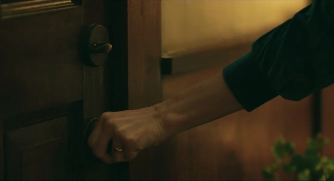 Haunter (2013) movie still: closeup — A wooden interior door features a classic brass handle and a matching lock plate. A person wearing a teal…