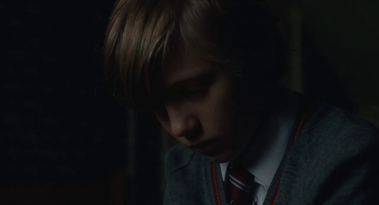 The Nest (2020) movie still: closeup — A dimly lit space features a student wearing a school uniform. The boy looks downward while sitting in a…