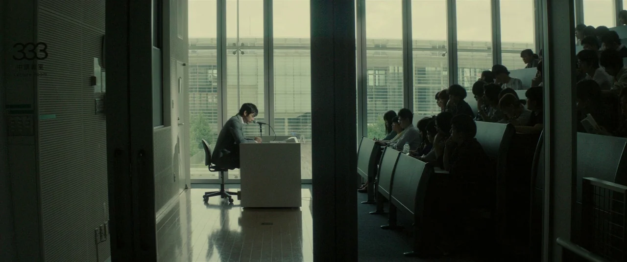 Creepy (2016) movie still: wide — A modern university lecture room with floor-to-ceiling windows overlooking an outdoor area. A male professor sits…