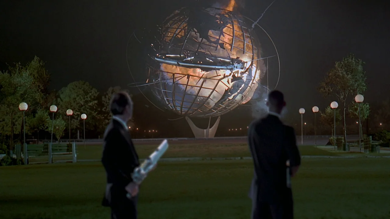 Men In Black (1997) movie still: wide — A nighttime city park features a large metallic globe monument enveloped in flames. Two men in dark suits…