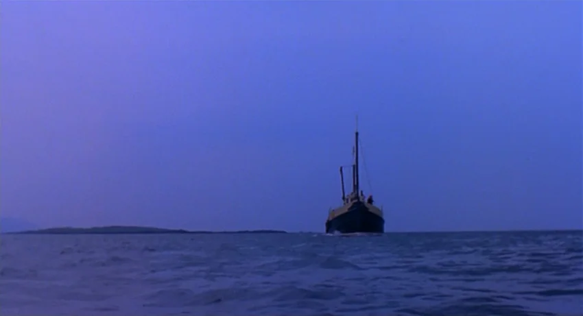 The Secret Of Roan Inish (1994) movie still: wide — A small fishing boat travels across a calm sea under a dark blue sky. A low landmass is visible…
