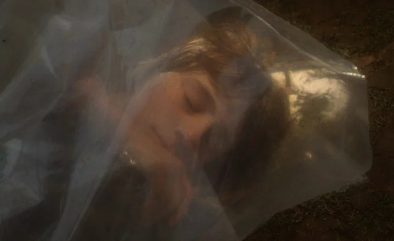 Sweet Movie (1974) movie still: closeup — A person lies on the ground, covered by a sheet of clear plastic. The background is dark earth with…