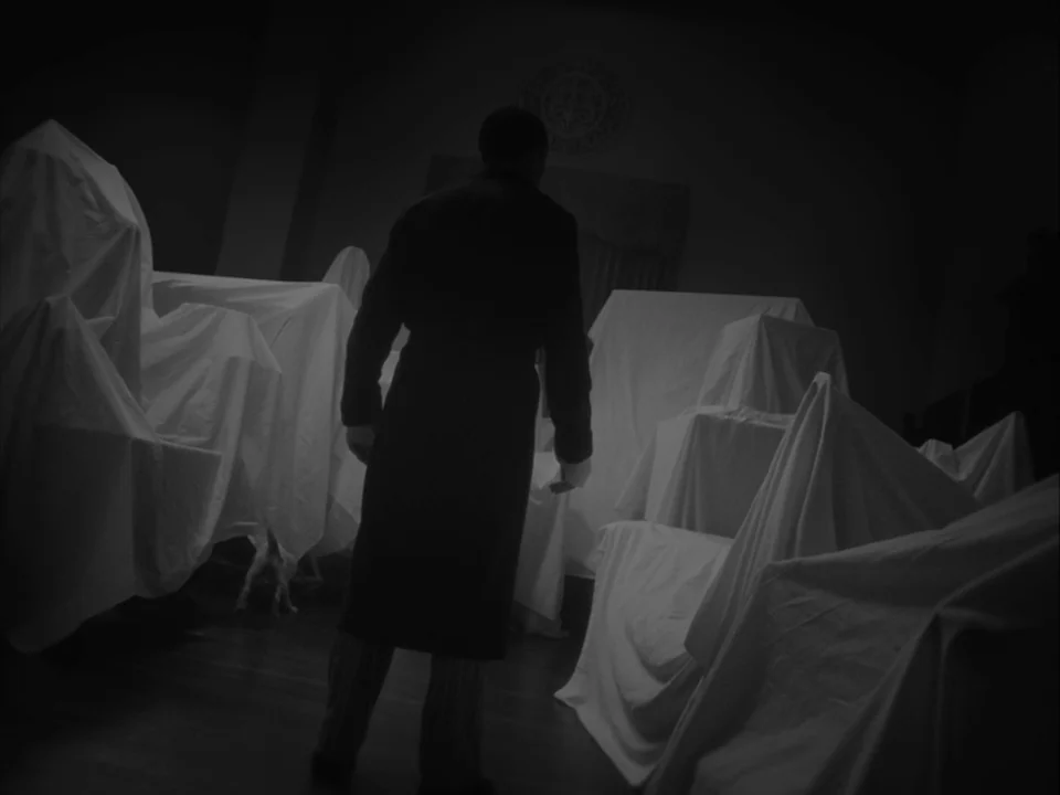 The Artist (2011) movie still: medium — A dark, cavernous space is filled with furniture hidden under white dust cloths. A man stands in the center…