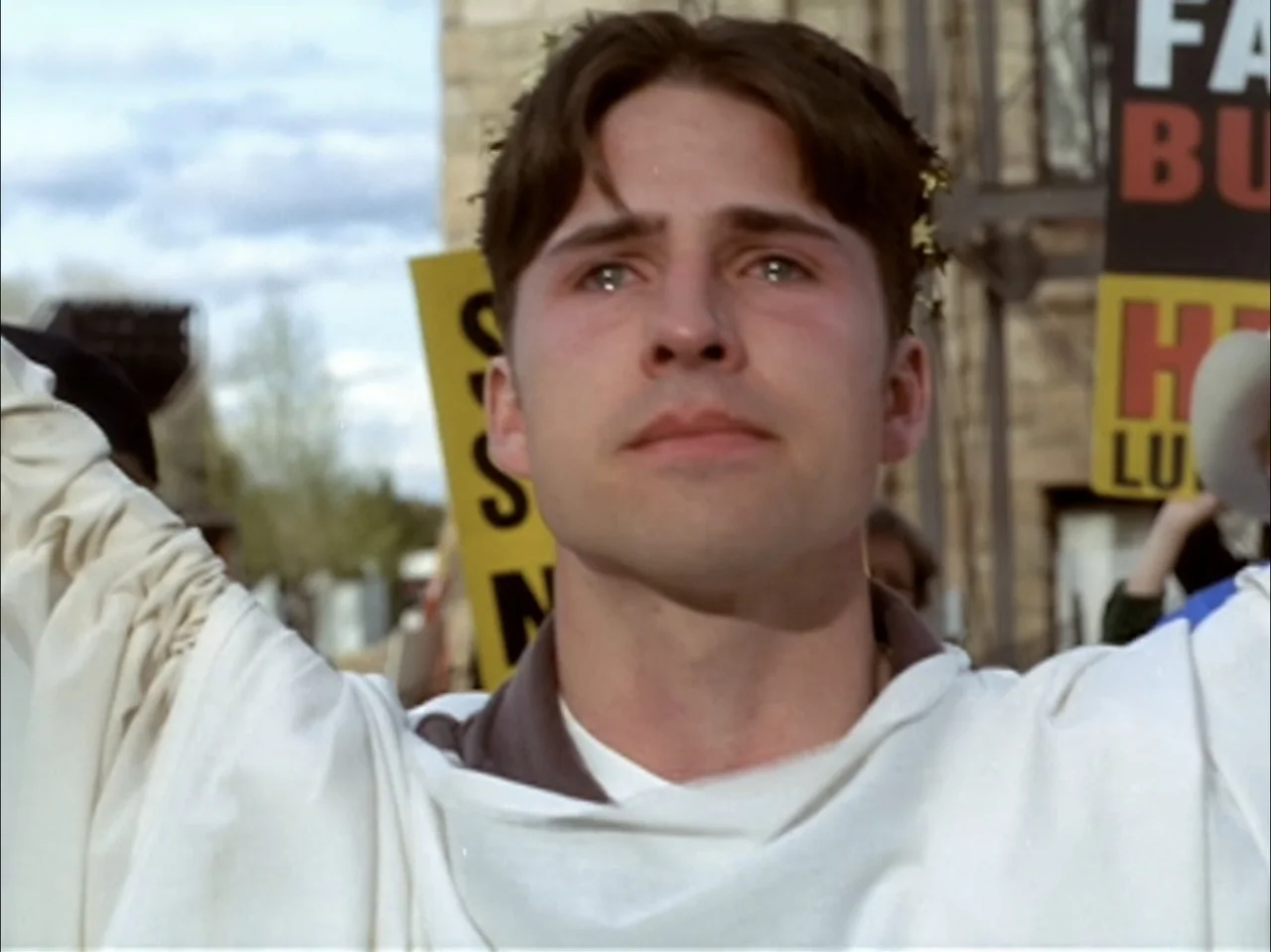 The Laramie Project (2020) movie still: closeup — A man in a white robe stands before a crowd holding signs. He raises his arms with a focused…
