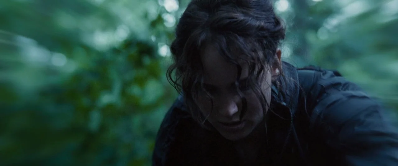The Hunger Games (2012) movie still: closeup — A woman is positioned amidst dense, blurred greenery in a natural outdoor environment. She leans…
