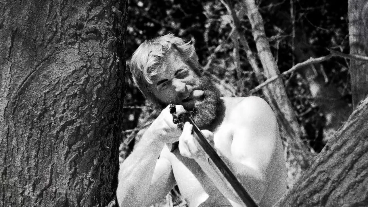 Hundreds of Beavers (2022) movie still: medium — A dense patch of woods with rough textured tree trunks framing the subject. A shirtless bearded man…