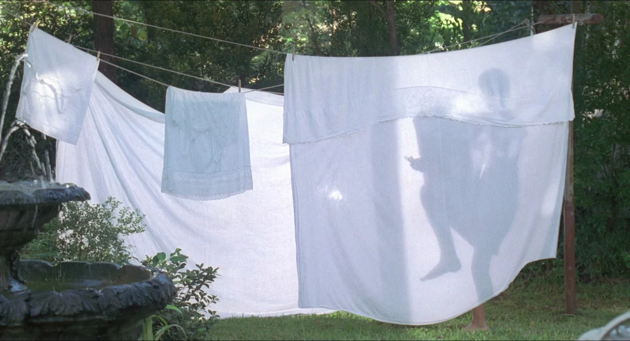 Lolita (1997) movie still: wide — White laundry hangs on a line in a sunlit outdoor green yard. A dark silhouette of a person appears through the…