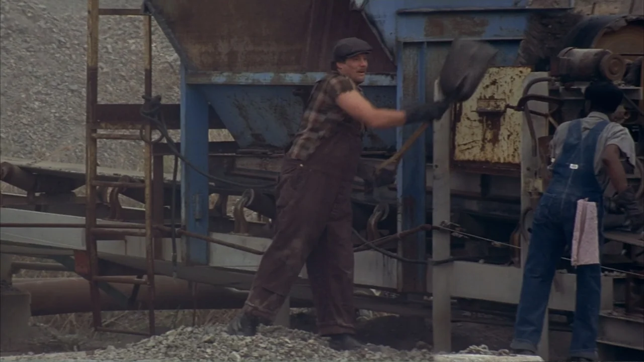 The Notebook (2004) movie still: fullbody — A dusty outdoor quarry features a large blue industrial metal sorting and crushing machine. Two laborers…