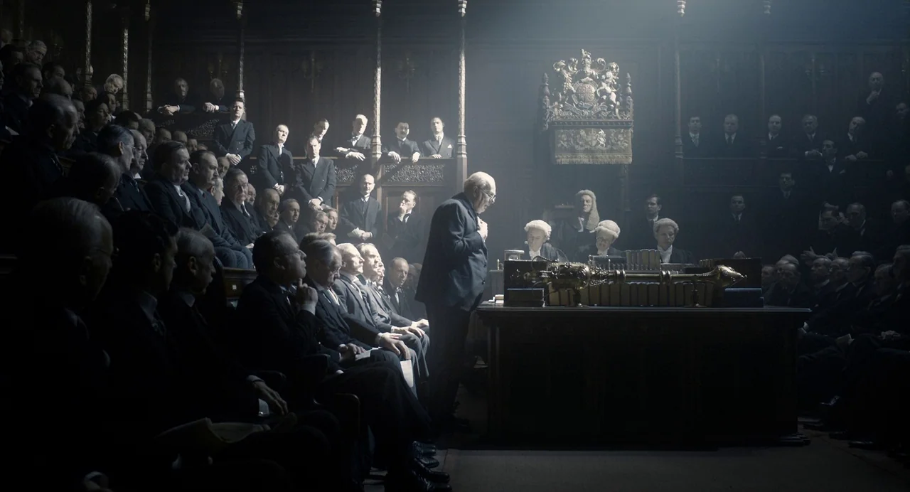 Darkest Hour (2017) movie still: wide — A dark, wood-paneled legislative hall features rows of seated men in formal suits. A lone man stands in a…