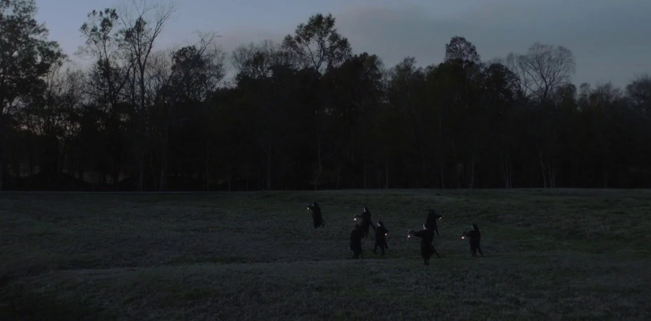 Darlin’ (2019) movie still: wide — A vast, open grass field is framed by a dense line of silhouetted trees. Several figures wearing dark habits…