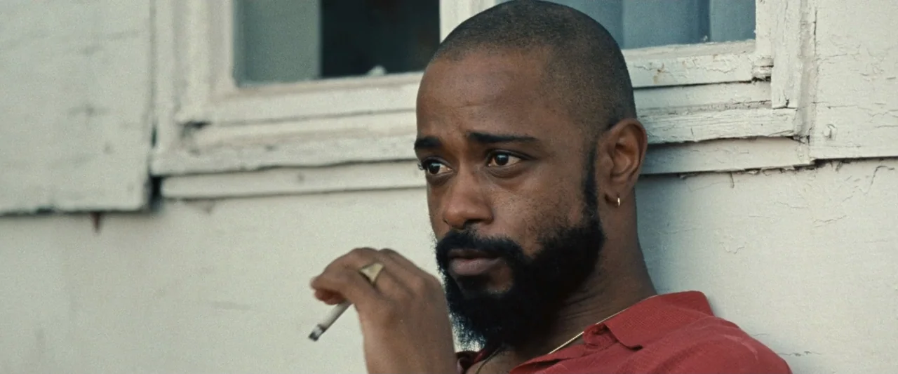 Roofman (2025) movie still: closeup — A man stands before a weathered white painted wall with chipped edges. The man holds a lit cigarette while…