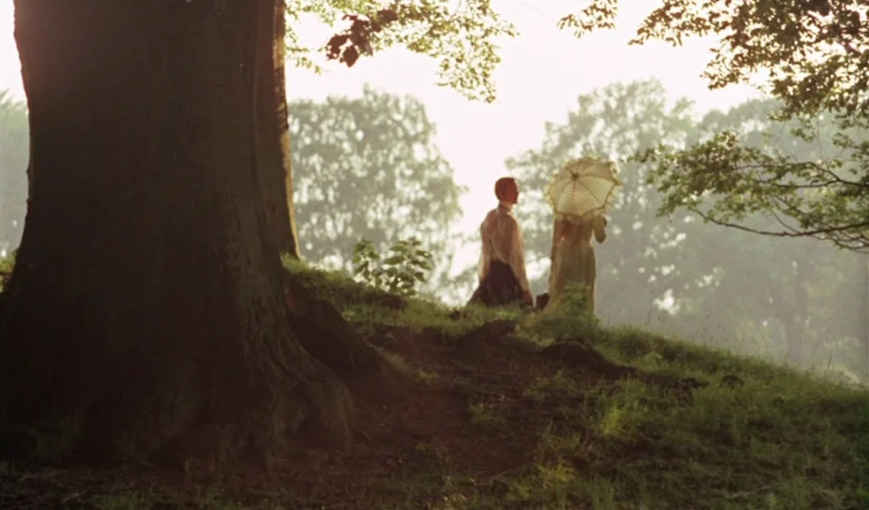 Elvira Madigan (1967) movie still: wide — A grassy hill with a large tree in the foreground under bright sunlight. A man and a woman in period…