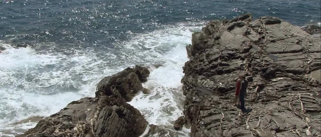 Genova (2008) movie still: wide — A rugged rocky coastline meets deep blue ocean water with white crashing waves. A man and a woman stand together…